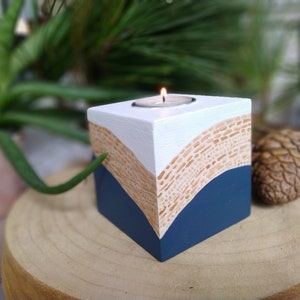 Peut inclure: Un bougeoir en bois blanc, bleu et beige avec une bougie chauffe-plat au centre. Le bougeoir a un design géométrique avec des lignes courbes et une surface texturée.