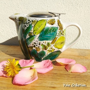 Può includere: Una teiera in ceramica bianca con un coperchio in acciaio inossidabile e un design floreale verde e giallo. La teiera ha un manico e un beccuccio. La teiera è appoggiata su una superficie di legno con petali di rosa rosa.