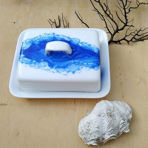 Può includere: Un porta burro in ceramica bianca con un design marmoreo blu e bianco. Il porta burro ha un coperchio con un manico.