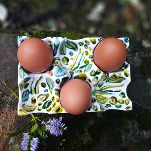 May include: A white ceramic egg holder with a green, yellow, and blue floral design. The holder has four compartments and is filled with brown eggs.