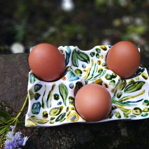 May include: A white ceramic egg holder with a green, blue, and yellow floral pattern. Three brown eggs are sitting in the holder.