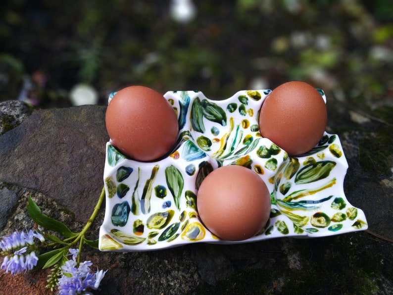 May include: A white ceramic egg holder with a green, blue, and yellow floral pattern. Three brown eggs are sitting in the holder.