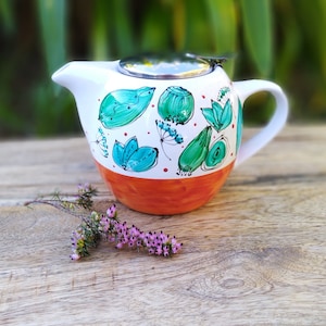 May include: A white teapot with a stainless steel lid and a handle. The teapot is decorated with a hand-painted design of green leaves and red dots on a white background. The bottom of the teapot is orange.