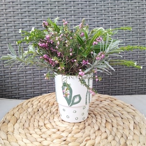 Peut inclure: Un pot en céramique blanche avec un motif floral vert et blanc, rempli de fleurs roses et blanches. Le pot est posé sur un tapis de paille tressé.