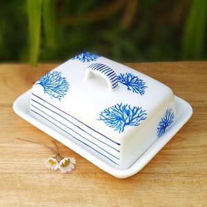 Può includere: Un porta burro in ceramica bianca con un design a corallo blu e bianco. Il porta burro ha un coperchio con manico e una base abbinata.