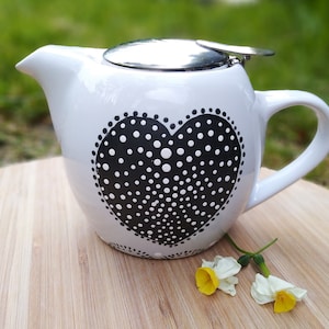 Peut inclure: Théière en céramique blanche avec un motif de cœur à pois noirs et un couvercle en acier inoxydable. La théière a une poignée et un bec verseur. La théière est posée sur une surface en bois.