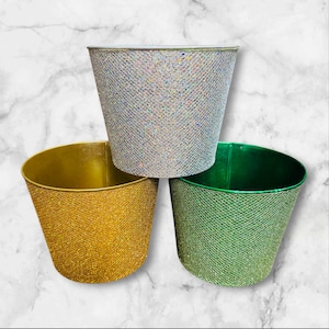 May include: Three decorative metal buckets in gold, silver, and green. Each bucket has a textured, glittery finish. The buckets are arranged in a triangular formation, with the gold bucket at the bottom left.