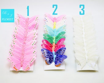 May include: Three sets of butterfly embellishments. The first set is pink with gold glitter dots. The second set is a rainbow of colors, including blue, green, pink, and purple. The third set is white and sheer. Each set is on a white card with a number 1, 2, or 3 printed on it.