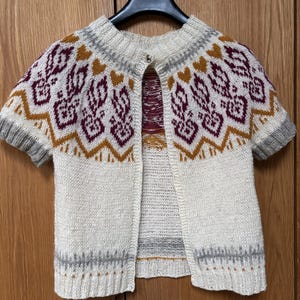 Puede incluir: Rebeca de manga corta color crema con cierre de botón. El suéter presenta un patrón geométrico en burdeos, amarillo mostaza y gris. El diseño incluye formas de diamantes y corazones, con ribete gris en las mangas y el cuello.