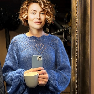Puede incluir: Una mujer lleva un suéter azul de punto con un delicado patrón de encaje. Está sosteniendo una taza de cerámica blanca y una funda de teléfono azul con una lente de cámara visible.