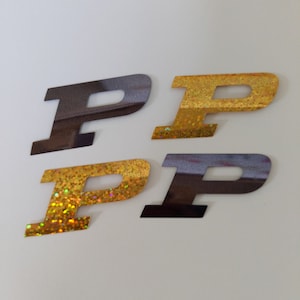 May include: Four letter "P" shaped stickers. Two are gold with glitter, and two are a dark brown color. The stickers are on a white background. The stickers are likely for crafts or scrapbooking.