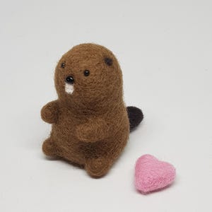 May include: A brown felt beaver with black eyes and a white nose, sitting next to a pink felt heart.