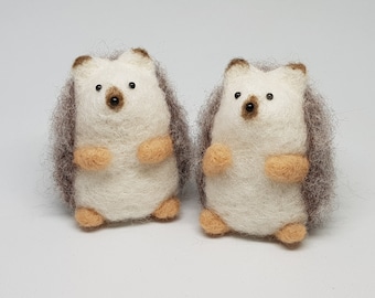 May include: Two small, white needle felted hedgehogs with brown noses and black eyes. The hedgehogs have brown fur around their faces and are standing on a white surface.