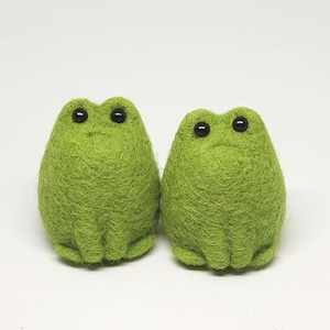 May include: Two handmade, green felt frog figurines with black bead eyes. The frogs are side-by-side, with a textured, fuzzy appearance. The background is plain white.