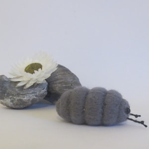 Pu&ograve; includere: Un millepiedi di feltro grigio con gambe nere, accanto a una roccia grigia con un fiore bianco in cima.