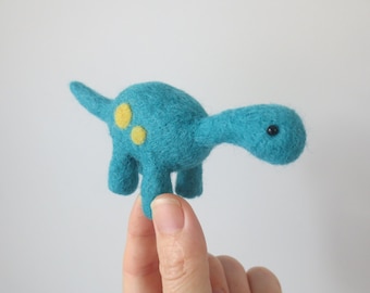 May include: A small, handmade felt dinosaur in a vibrant teal color. The dinosaur has a long neck, a small tail, and yellow spots. The eyes are small black beads. A charming, whimsical toy.
