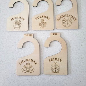 May include: Five wooden hangers with days of the week written on them. Each hanger has a different floral design.