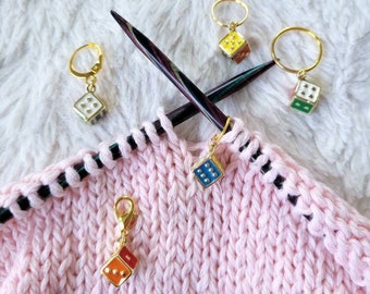 Può includere: Quattro marcatori di punto a forma di dado colorati con anelli e chiusure dorate. I marcatori sono su un tessuto a maglia rosa con strisce nere. Due ferri da maglia sono visibili sullo sfondo.
