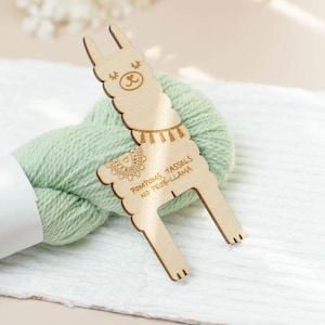 May include: A wooden llama-shaped pompom and tassel maker with the text "POMPOMS, TASSELS, NO PROB-LLAMA" is shown next to a ball of light green yarn. The maker has a decorative design and is made of light-coloured wood.