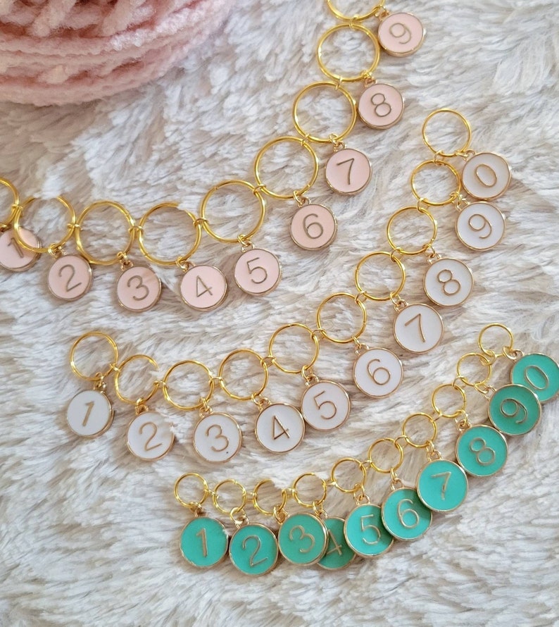 May include: Three sets of gold stitch markers with numbered charms. The first set has pink charms with gold numbers 1-9. The second set has white charms with gold numbers 1-9. The third set has turquoise charms with gold numbers 1-9.