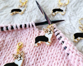 May include: Four gold stitch markers with a black and white corgi design. The markers are attached to a pink knitted fabric.