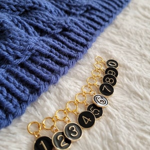 May include: A set of ten gold stitch markers with black round tags numbered 1 through 8 and 0. The tags have gold numbers.