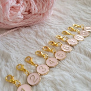 Könnte beinhalten: Zehn goldfarbene Maschenmarkierer mit runden rosa Scheiben, die von 1 bis 10 nummeriert sind. Die Markierer haben einen federbelasteten Verschluss.