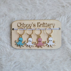 May include: Four octopus-shaped stitch markers in blue, white, and pink with gold-colored rings, displayed on a wooden tag. The tag has the text "Chippy's Knittery" engraved on it.