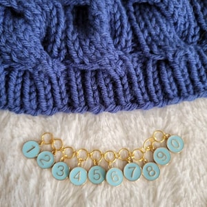 May include: A set of ten gold stitch markers with blue enamel and white numbers 1 through 10. The markers are linked together in a chain.