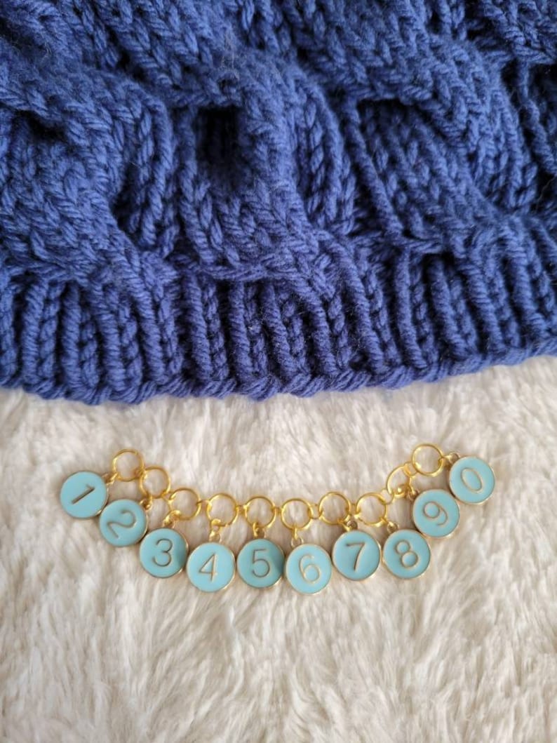May include: A set of ten gold stitch markers with blue enamel and white numbers 1 through 10. The markers are linked together in a chain.