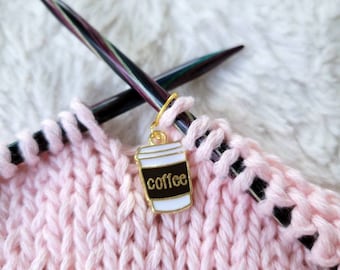 May include: A pink knitted fabric with black knitting needles and a gold coffee cup stitch marker. The coffee cup has the word "coffee" printed on it.