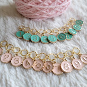 May include: Two sets of gold chain stitch markers with numbered charms. One set has teal charms with gold numbers, the other has pink charms with gold numbers.