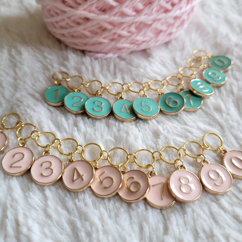 May include: Two sets of gold chain stitch markers with numbered charms. One set has teal charms with gold numbers, the other has pink charms with gold numbers.