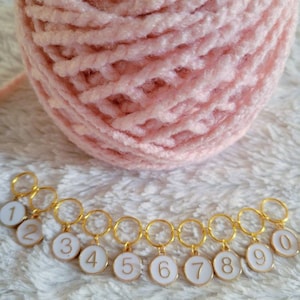 May include: A set of ten gold-colored stitch markers with white enamel and black numbers 1 through 10. The markers are linked together in a chain.