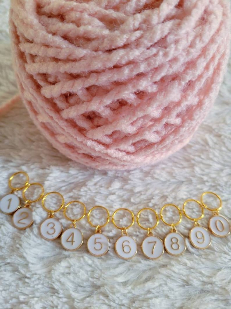 May include: A set of ten gold-colored stitch markers with white enamel and black numbers 1 through 10. The markers are linked together in a chain.