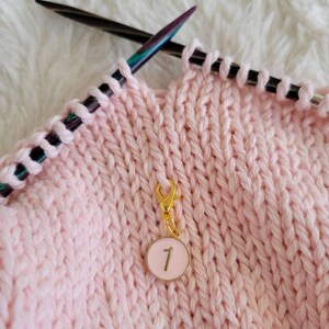 May include: A pink knitting project with a gold stitch marker with the number "1" on a white background.