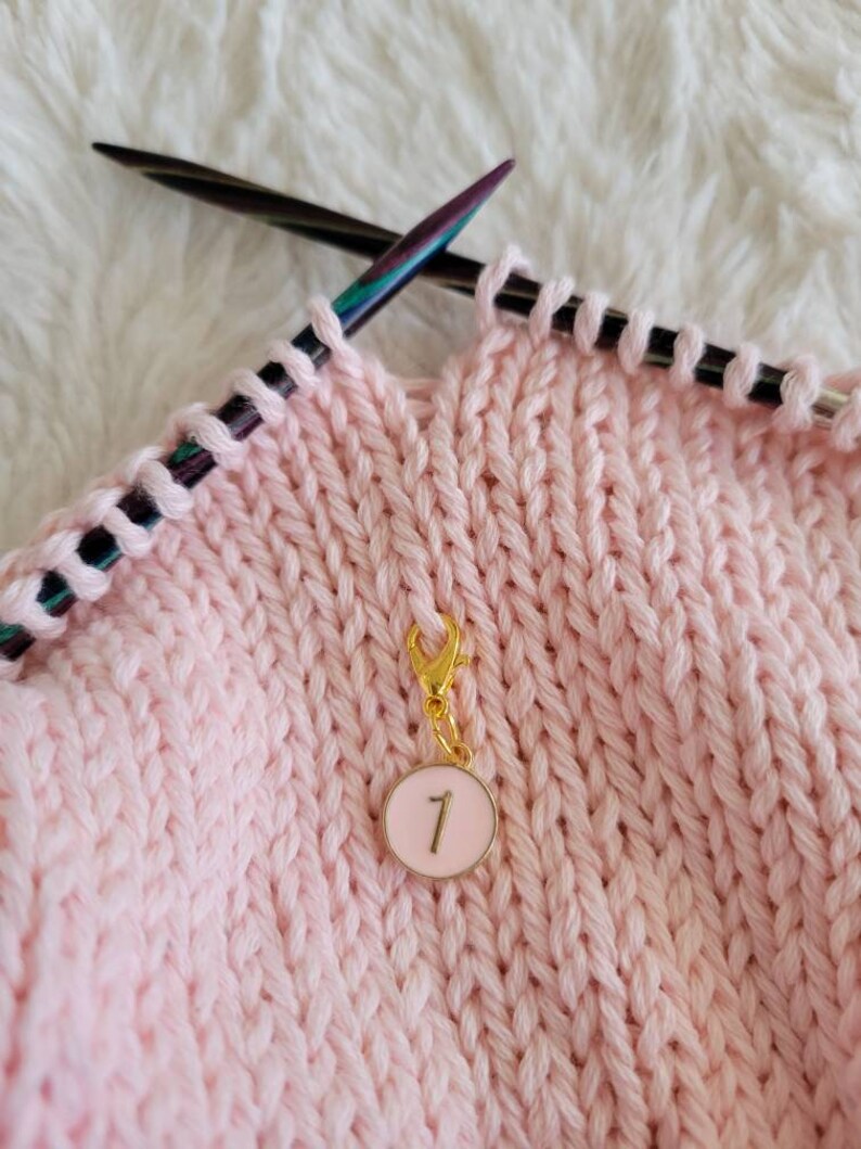 May include: A pink knitting project with a gold stitch marker with the number "1" on a white background.