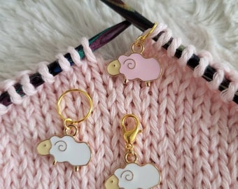 May include: Three gold sheep stitch markers on a pink knitted fabric. Two of the sheep are white with gold accents and one is pink with gold accents.
