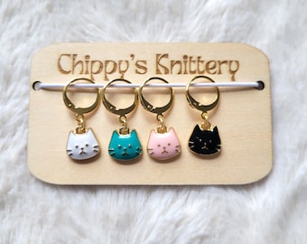 May include: Set of four gold cat stitch markers with white, teal, pink, and black enamel. The markers are displayed on a wooden board with the text "Chippy's Knittery".