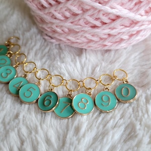 May include: A set of ten gold and turquoise stitch markers with numbers 1 through 10. The markers are connected by a gold chain.