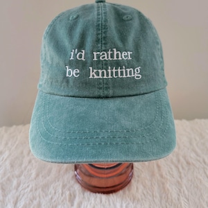 May include: A teal baseball cap with the embroidered text "i'd rather be knitting" in white. The cap has a curved brim and a fabric strap closure. The hat is made of a soft, washed cotton material.