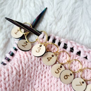 Könnte beinhalten: Nahaufnahme eines rosa gestrickten Stoffes mit goldenen Maschenmarkierern, die von 1 bis 8 nummeriert sind. Die Markierer sind an einer Stricknadel befestigt.