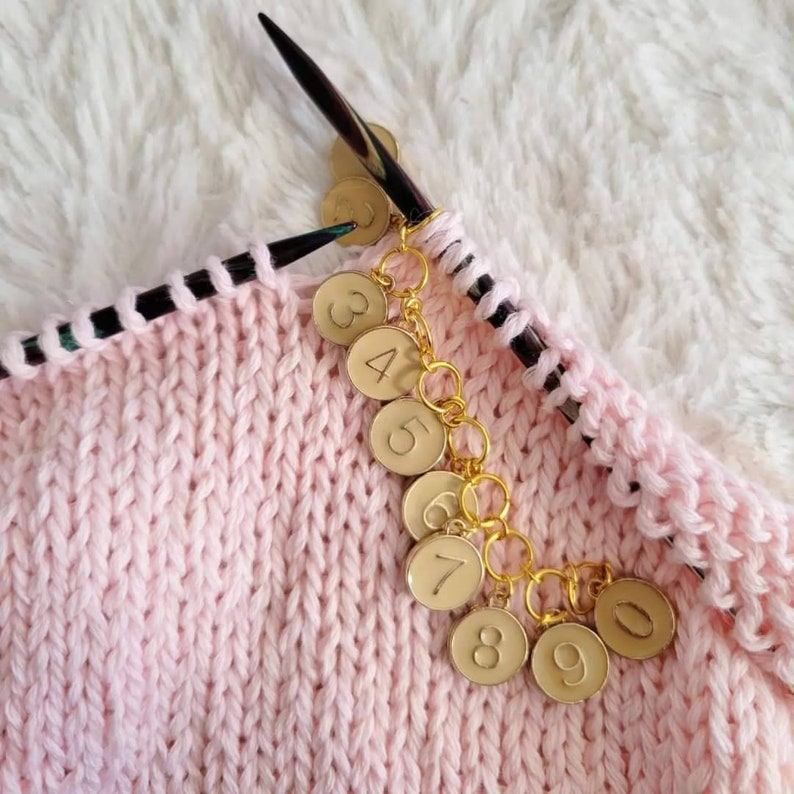 May include: A close-up of a pink knitted fabric with a gold chain of numbered stitch markers. The numbers on the markers are 3, 4, 5, 6, 7, 8, 9, and 0.