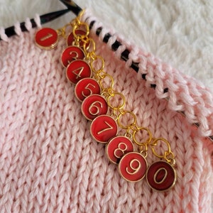 May include: A set of ten red and gold stitch markers with numbers 1 through 10. The markers are attached to a chain and are used to mark rows or stitches in knitting.