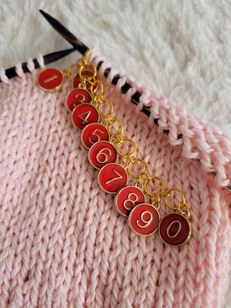 May include: A set of ten red and gold stitch markers with numbers 1 through 10. The markers are attached to a chain and are used to mark rows or stitches in knitting.