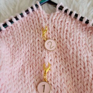 May include: Two pink and gold stitch markers with numbers 1 and 2 on them. The markers are attached to a pink knitted fabric.