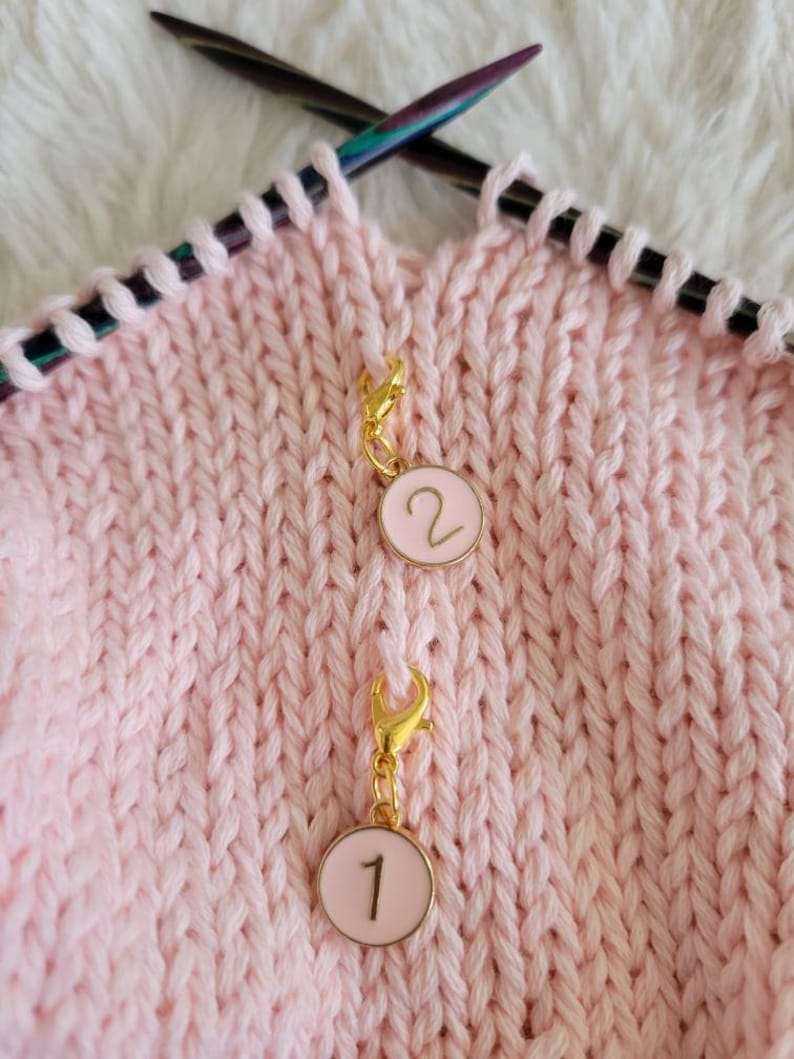 May include: Two pink and gold stitch markers with numbers 1 and 2 on them. The markers are attached to a pink knitted fabric.