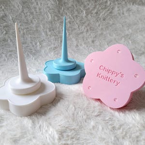 May include: Three flower-shaped bases for knitting needles. The bases are white, light blue, and pink. The pink base has the text "Chippy's Knittery" on it.