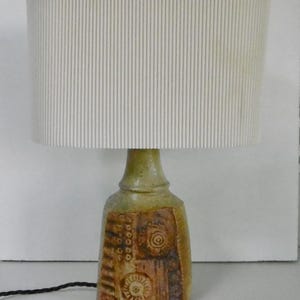 May include: A table lamp featuring a cylindrical, off-white, vertically ribbed lampshade atop a hand-thrown ceramic base. The base has a mottled green and brown glaze with embossed circular and linear patterns and a black electrical cord.