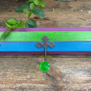 May include: A wooden wall-mounted coat rack with three hooks. The rack is painted in shades of purple, blue, and green. Each hook has a decorative metal finial and a green glass knob.